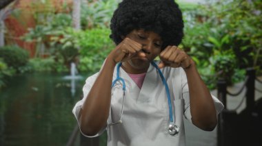 Steteskoplu önlük giyen kadın doktor yeşil bir parkta gölgelik bir gölette gözlerini ovuşturuyor..