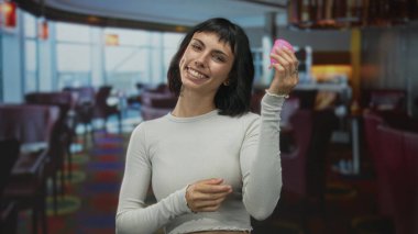 Çıplak elle pembe regl bardağı tutan ve restoranı işaret eden kadın; sürdürülebilir dönem özgüveni.