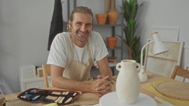 Önlük takan sarışın adam, güneş ışığı altındaki bir stüdyoda, ahşap bir masanın üzerinde seramik bir vazonun yanında dinleniyor..