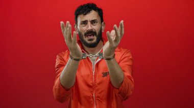 Portakal rengi hapishane tulumlu sakallı, elleri kelepçeli İspanyol bir adam, canlı kırmızı bir arka plana karşı duruyor, hapis ve kısıtlama sahnesini betimliyor..