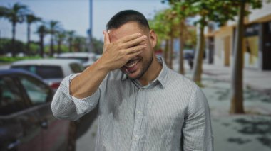 Adam gülümser ve elleri sokakta park halindeki arabalarla ve palmiye ağaçlarıyla kaplı gözlerle kapatır; utanç.