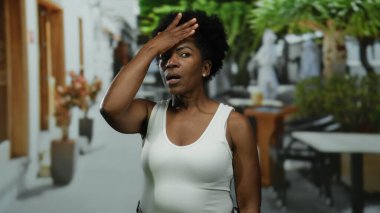 Şehir restoranındaki terasta endişeli bir kadın, dışarıda kolsuz gömlekle saksıların ve masaların arasında dikilirken düşünceli bir şekilde alnına dokunuyor..