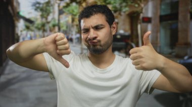 Şüpheci çatık kaşlı genç İspanyol adam kalabalık bir şehir sokağında baş parmağını kaldırdı ve baş parmağını kaldırdı; seçim kararsızlık.