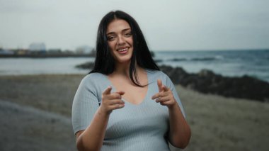 Uzun saçlı, gülümseyen ve kumsaldaki kameraya bakan bulutlu bir günde arka planda deniz olan genç bir kadın kendinden emin ve kıvrımlı vücudunu sergiliyor..