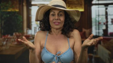Mavi bikinili ve güneş şapkalı İspanyol kadın şık bir restoran ortamında el kol hareketleri yapıyor. Günlük ama kendine güveni tam tavrını sergiliyor..