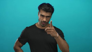 Siyah tişörtlü genç adam stüdyoda kameraya ciddi bir ifade ve dik duruşla işaret ediyor. Kararlılık..