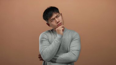 Gri gömlekli genç adam kahverengi bir arka plan üzerinde düşünceli bir ifade ile, bir stüdyo ortamında tefekkür ve içgözlem örneği..