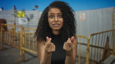 Genç bir kadın ellerini sarı bir bariyerin yanında birbirine bastırır ve bina girişini açık havada yapar; çaresizlik.