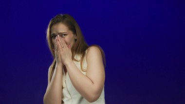 Mavi bir stüdyoda beyaz bir elbise giyen korkmuş yüz ifadesi olan beyaz bir kadın, korku..