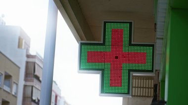 Güneşli bir şehir sokağında, arka planda ilaç hizmetlerini gösteren binalar bulunan bir eczane kapısı..