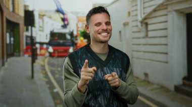 Sakallı genç bir adam, kenar mahallede kendine güvenerek gülümsüyor. Açık havada moda anlayışı sergiliyor. Olumlu hareketler yapıyor..