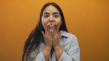 Şaşırmış ifadeye sahip genç bir kadın turuncu arka planda ağzı çizgili bir gömlekle kaplıyor. Neşeli ve anlamlı bir şok ve heyecan anı yaşıyor..