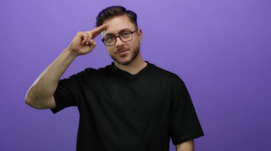 Sakallı, gözlüklü, parmakları mor arka planda selam veren, günlük siyah gömlek giyen, stüdyo ortamında dostane ve cana yakın bir jest yapan genç bir adam..