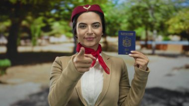 Genç İspanyol uçuş görevlisi üniformalı gülümseyip işaret ederken Birleşik Arap Emirlikleri pasaportunu açık mavi gökyüzünün altında tutuyor..