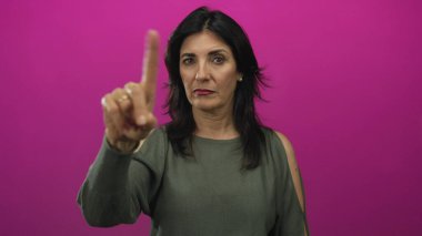 Zeytin rengi kazaklı orta yaşlı kadın pembe stüdyoda parmağıyla kameraya işaret ediyor, inci küpeler takıyor;.