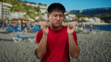 Kırmızı gömlekli genç adam, güneşli sahilde şemsiyeli ve açık mavi gökyüzüyle rahatsızlığını dile getiriyor. Okyanus kenarında deniz kenarında bir yaz atmosferi olduğunu gösteriyor..