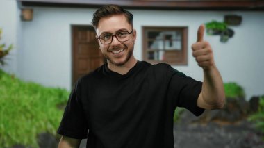 Sakallı genç bir adam şehir sokaklarında gözlüklü ve siyah tişörtlü, şehir ortamında karışık duygular ifade eden bir sokak sahnesinde başparmak kaldırıyor..