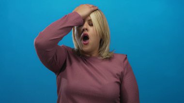 Mavi bir stüdyo duvarına karşı eli alnında ve gözleri kapalı genç sarışın kadın şaşkınlık.