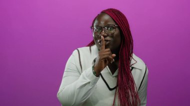 Gözlük ve beyaz süveter giyen, soyutlanmış pembe bir arka plana karşı sessiz kalmak için el kol hareketi yapan Afrikalı Amerikalı bir kadın..