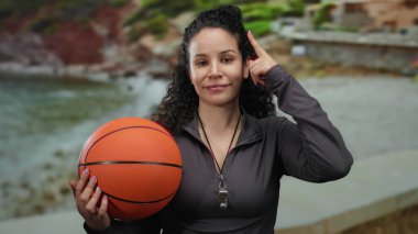 Plaj sahilinde basketbol oynayan bir kadın, boynunda ıslık çalan, açık gökyüzünün altında rahat ve düşünceli bir ifade sergiliyor..