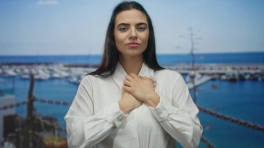 Elleri göğsünde, arka planda teknelerle limanda duran, beyaz gömlek giyen, açık denizde duygusal ve yansıtıcı bir an yaşayan bir kadın..