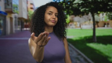 Bulanık binalar ve ağaçlarla şehir caddesinde kameraya doğru elini açık tutan Latin kökenli genç bir kadın..