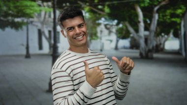 Genç adam arka planda ağaçlar ve binalar olan bir caddede başparmak kaldırıyor, çizgili bir gömlek giyiyor ve kendine güvenerek gülümsüyor, pozitif tavır sergiliyor..