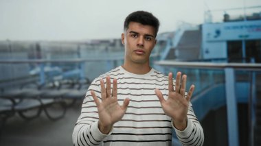 Bir yolcu gemisinin güvertesinde, elleri havada, çizgili gömlek giymiş, arka planda bulanık oturma ve gemi korkulukları olan bir adam..