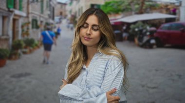 Çapraz kollu genç bir kadın tarihi bir köyün kaldırım taşı sokakta aşağı bakıyor; sakin barış.