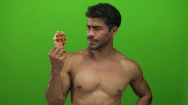 Tişörtsüz İspanyol adam bir waffle tutuyor ve bir stüdyoda yeşil bir duvara karşı burnunu kırıştırıyor..
