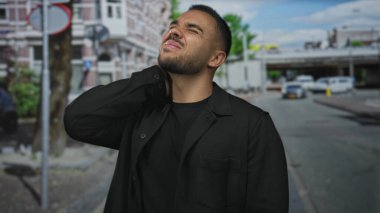 Siyah ceketli adam bulutlu gökyüzü altında şehir binalarının ortasında park halindeki arabaların yanında boynunu tutuyor;.