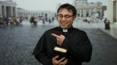 Papaz kıyafeti giymiş Asyalı bir rahip elinde kitap tutuyordu ve arka planda turistlerin bulunduğu Vatikan 'ın St. Peter Meydanı' nda gülümsüyordu..