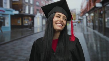 Mezuniyet şapkalı İspanyol kadın, şehir sokaklarında canlı kentsel çevrede elde edilen başarıları kutlarken, arka planda binalar ve dükkanlar ile gülümsüyor.