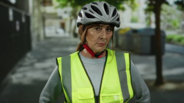 Kasklı ve yansıtıcı yelek giyen İspanyol kıdemli kadın bir şehir caddesinde, gözleri kapalı duruyor, şehir ortamında konsantrasyon veya meditasyon ima ediyor..