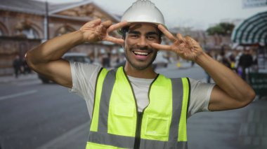 Beyaz kasklı ve yansıtıcı yelekli adam şehir sokaklarında gülümserken iki parmağını da öne doğru gösteriyor..