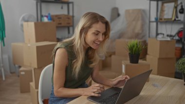 Yeni evinde dizüstü bilgisayar kullanan bir kadın oturma odasındaki kutularla çevrili genç, sarışın, beyaz bir kadını yeni taşınmış bir apartmanda sergiliyor..