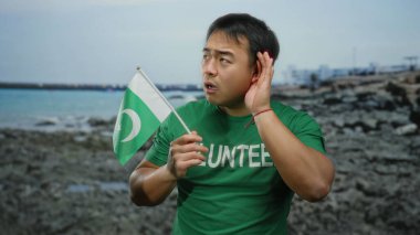 Plajda elinde Pakistan bayrağı olan genç bir adam dikkatle dinliyormuş gibi kulak kesiliyor, yeşil gönüllü gömleği giyiyor, deniz kenarındaki kültürel çeşitliliği temsil ediyor..