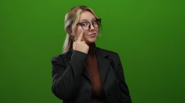 Siyah ceketli, yeşil arka planda gözlüklü, parlak renklerde bir stüdyo ortamında kendine güveni ve pozitifliği gösteren bir kadın..