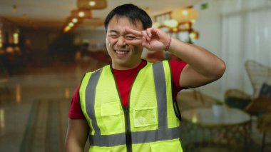 Otelin içinde güvenlik yeleği giyen bir adam iyi ışıklandırılmış arka planda güvenle gülümserken aynı zamanda modern iç mekan atmosferini ve günlük kıyafetleri ön plana çıkarıyor..