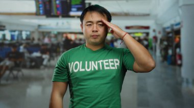 Havaalanı terminalinde yeşil gönüllü gömleği giyen, arka planda bulanık yolcu ve kalkış ekranları olan genç bir adam seyahat ve yardım bağlamını çağrıştırıyor..