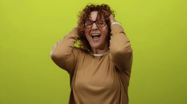 Sarı arka planda hayal kırıklığına uğramış, orta yaşlı, kıvırcık esmer saçlı, rahat süveter ve gözlük giyen, elleri umutsuzluk içinde olan bir kadın..