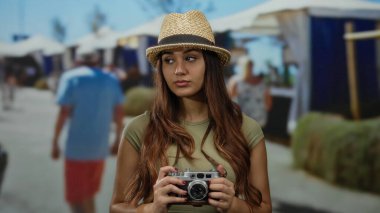 Şapkalı bir kadın elinde kamera, canlı bir açık hava pazarında dikiliyor. Sokakta insanlarla ve dükkanlarla dolu anları yakalıyor..