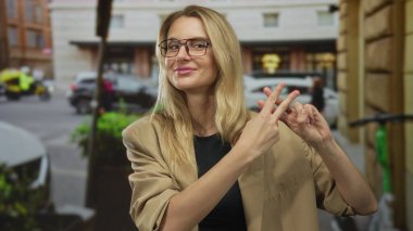 Meşgul şehir caddesinde hashtag jesti yapan bir kadın sarışın gözlüklü ve açık havada günlük bej ceketli şehir yaşamını sergiliyor..
