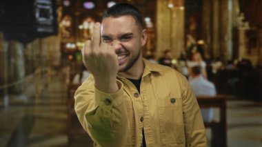 Genç İspanyol adam orta parmağını sallıyor ve kilise binasının içinde ahşap sıralar ve avizelerle yumruklarını sıkıyor..