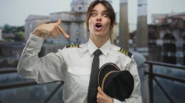 Üniformalı kadın pilot, açık şehir ortamında antik Roma harabelerinin arka planına gülümseyen gururu ve kimliğini vurgulayan kendini işaret ediyor..