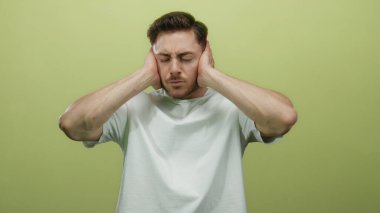 Sakallı genç adam sarı arka planda yalnız. Kulakları kulaklarla kapatıyor. Stres ve hayal kırıklığını ifade ediyor..