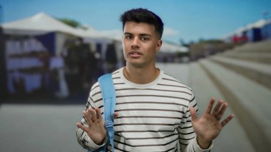 Şehir caddesinde mavi sırt çantalı genç bir adam açık havada çizgili gömlek giyiyor güneşli bir günde düşünceli görünüyor satıcıların ve basamakların bulanık geçmişiyle.