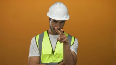 Beyaz şapka ve yansıtıcı yelek giyen genç İspanyol adam ve mühendis stüdyoda kameraya işaret parmağıyla işaret veriyor..
