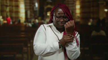 Kilisenin içinde bileğini tutan Afrikalı Amerikalı bir kadın vaaz sırasında acı çekiyor. Arka planda bulanık bir cemaat var. Gözlük ve örgü takıyor..