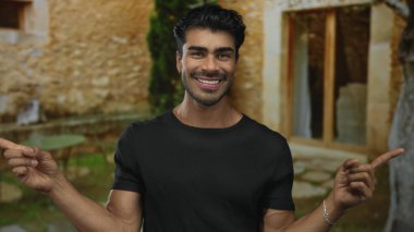 Kırsal bina girişini işaret eden parmaklarıyla gülümseyen bir adam taş duvara karşı bilezik takıyor. Hoş geldiniz..